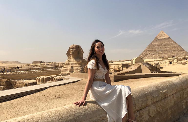 A female wearing white dress, sitting with a backdrop of Historic Great Sphinx and Pyramids of Giza in Egypt.