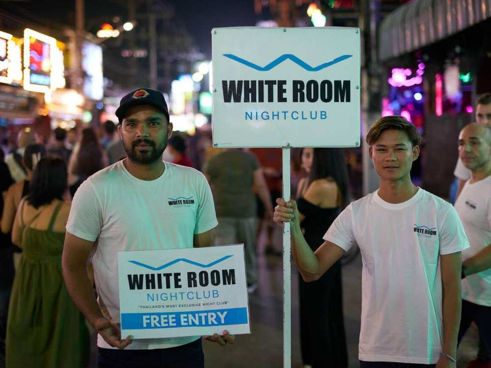 Two men holding advertisement posters on Patong Beach’s Bangla Walking Street at night, surrounded by neon lights and a busy nightlife crowd.