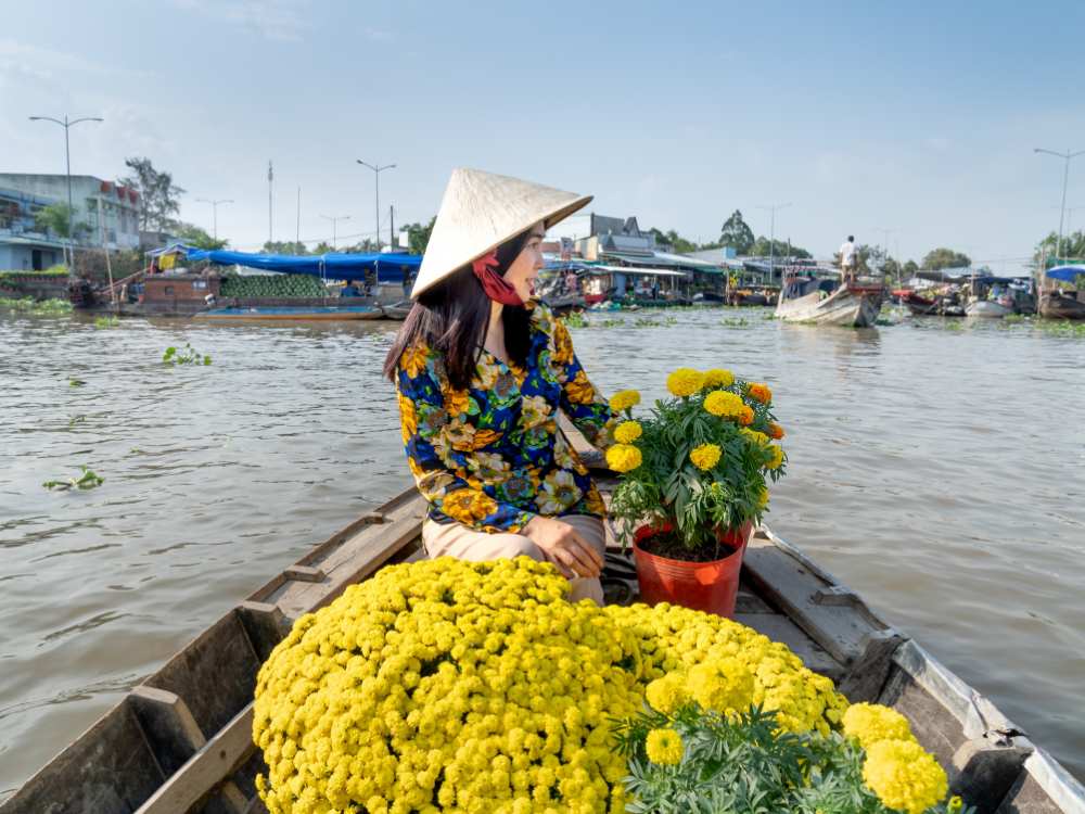 A woman wearing a bamboo hat, sitting in a boat with yellow flowers in front of her in Mekong delta of Vietnam